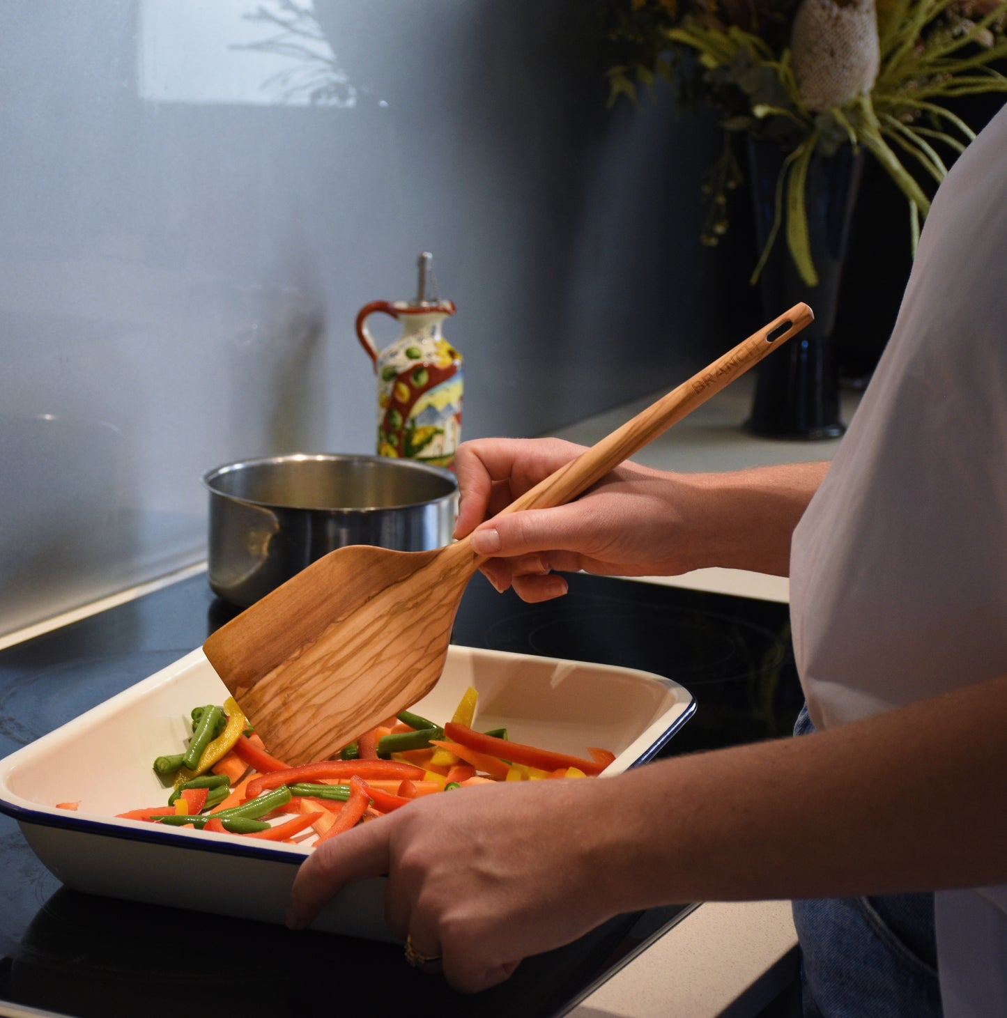 Person stirring vegetables in a baking dish with a wooden spoon, kitchen setting.